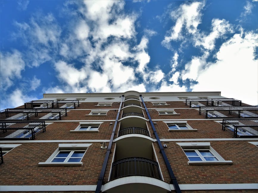 An exterior view of a modern multi-storey residential building featuring glass balcony railings, large windows, and a combination of white and beige facades. The building has multiple floors with visi