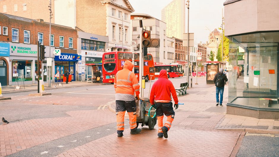 Two street cleaners in bright orange and red reflective uniforms are performing surface cleaning on a pedestrian pathway at Vicarage Road, Watford. They are pushing a cleaning cart with tools and hose